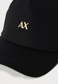 Melna beisbola cepure ar zelta metāla "AX" logotipu uz priekšējā paneļa, parādot detalizētu auduma tekstūru un šuvju. 