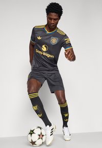 Γκρι ποδοσφαιρική φανέλα με χρυσές λεπτομέρειες, ταιριαστά σορτς και μαύρες κάλτσες με κίτρινες ρίγες. Ο παίκτης αλληλεπιδρά με μια άσπρη και μαύρη μπάλα ποδοσφαίρου.