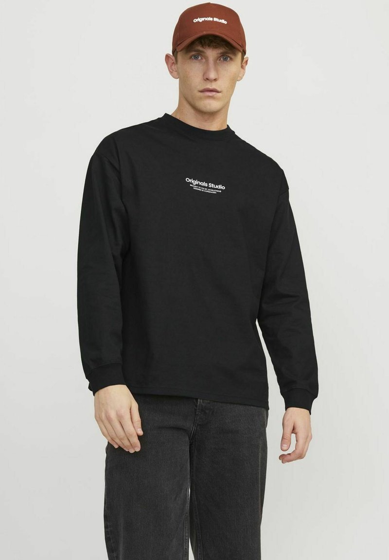 Svart långärmad t-shirt med rund halsringning och vit "Originals Studio" logotyp på bröstet, parat med en brun keps.