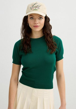 Mujer con cabello largo, castaño y ondulado, que lleva una gorra beige con texto floral, un suéter verde de manga corta y una falda plisada color crema sobre un fondo liso.