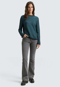 Pull texturé turquoise avec un col bateau, associé à un jean gris évasé et des chaussures plateformes noires. Le mannequin pose les mains dans les poches.