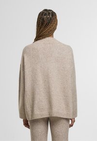 Béžový oversized svetr s texturovaným pletením, spadlými rameny, žebrovaným lemem a volným střihem. Zobrazeno zezadu.