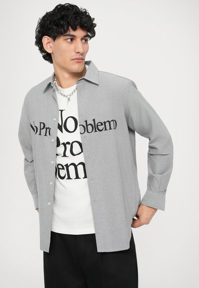 Grau karierte Hemd mit Kragen, mit fettem schwarzen Textdruck "Kein Problem". Getragen über einem weißen T-Shirt und schwarzen Hosen.