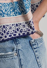 Top textil multicolor con patrones geométricos en azul, blanco y coral. Presenta un dobladillo en forma de ondas y detalles bordados. Llevado con jeans de mezclilla claros.