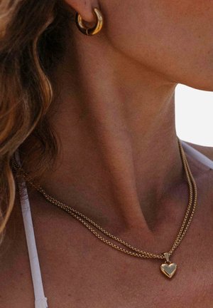 Nahaufnahme einer Frau, die goldene Creolen und eine doppelte Goldkette mit einem herzförmigen Anhänger trägt.