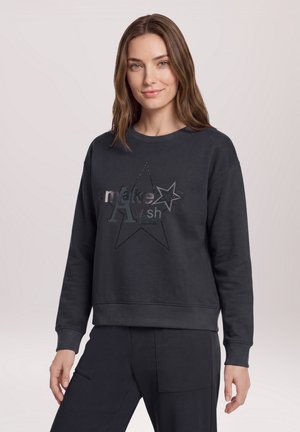 Femme aux cheveux châtain clair portant un sweat-shirt noir avec un motif étoile et les mots "make A wish" sur le devant.