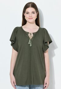 Olivi vihreä paita, jossa on lyhyet lepattelevat hihat, pyöreä pääntie ja pitsiyksityiskohtia. Edessä on nyöri, jossa on tupsut.