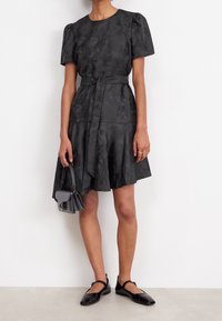 Robe noire texturée avec de courtes manches bouffantes, une ceinture à la taille et une jupe évasée. Associée à des chaussures plates noires et un petit sac à main.