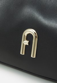 Juodas odinis rankinės modelis su blizgiu auksinės spalvos logotipo akcentu. Lygi tekstūra su kreivėmis formomis. Kompaktiška, elegantiška forma.