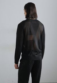 Schwarzer Pullover mit transparenter Einsätzen auf dem Rücken, kombiniert mit strukturierten schwarzen Hosen mit erhabenen Punkten. Einfaches, modernes Design.