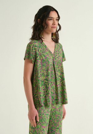 Donna che indossa una camicetta e pantaloni a maniche corte abbinati con motivo astratto verde e viola, con capelli lunghi e scuri e orecchini colorati con perline.