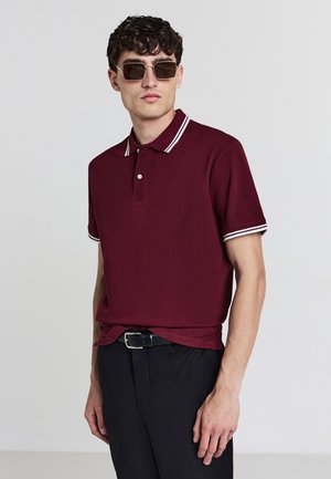 Polo burgundy avec col et poignets à rayures blanches. Texture lisse, patte de boutonnage, associé à un pantalon noir et des lunettes de soleil.