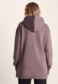 Oversized hoodie in muted mauve, featuring a large hood, long sleeves, and soft, textured fabric. The back has a relaxed fit and side slits.