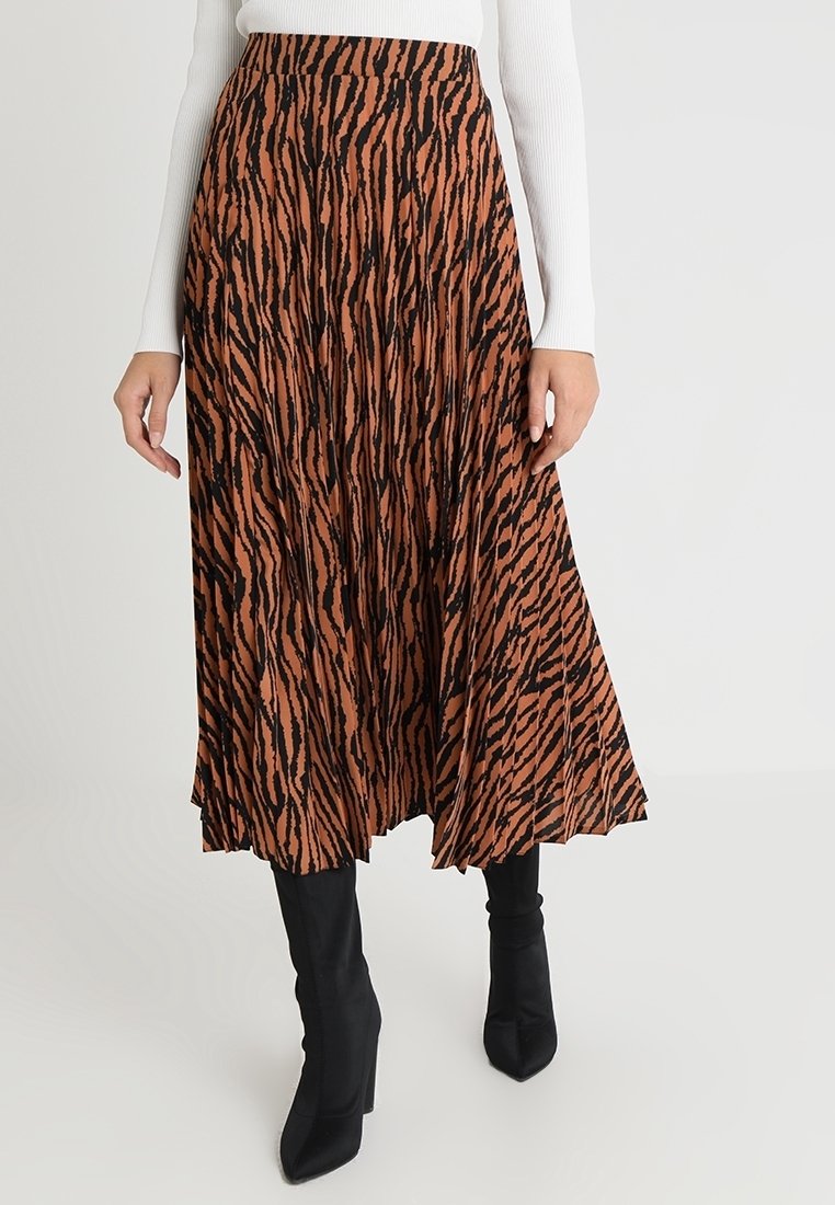 Femme portant une jupe midi plissée à rayures tigre marron et noir, des bottes à talons noires et un haut blanc à manches longues.
