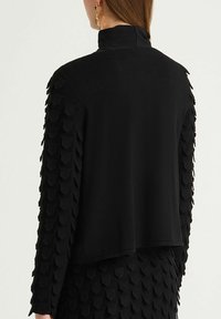 Haut noir à col montant, manches texturées avec détails superposés en forme d'écailles, et corps à finition lisse. Ourlet net.