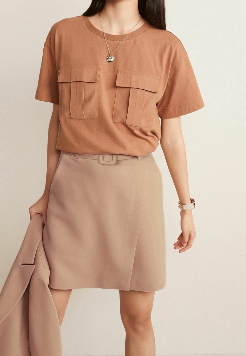 Femme portant une chemise à manches courtes couleur fauve avec deux poches poitrine, jupe beige ceinturée, tenant une veste assortie dans sa main gauche.