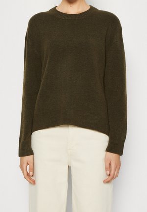 Dunkelgrüner Pullover mit rundem Ausschnitt, überschnittenen Schultern und gerippten Ärmeln sowie Saum. Weiche Textur, leicht lockere Passform. Kombiniert mit hellen Hosen.