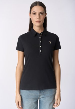 Donna con capelli lisci e scuri indossa una polo nera a maniche corte con abbottonatura e jeans azzurri, su uno sfondo uniforme.