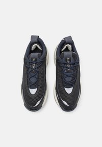 Melnas un tumši zila sieta kedas ar noapaļotu purngali, kurām ir teksturēta zole un pelēki akcenti. Pārsējo sprādze ir tumši zila; mēles daļā ir logotipa detaļa.