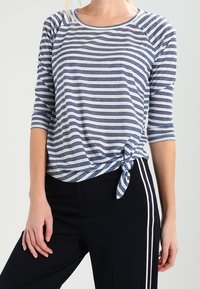 Kvinna klädd i en blå och vit randig skjorta med knut vid midjan, kombinerad med svarta byxor med vita sidostråkar.