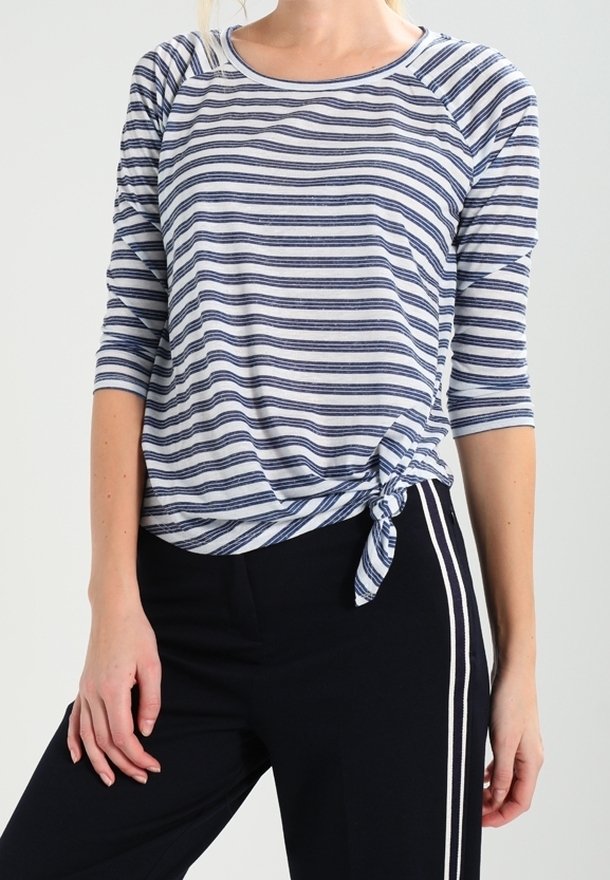 Kvinna klädd i en blå och vit randig skjorta med knut vid midjan, kombinerad med svarta byxor med vita sidostråkar.