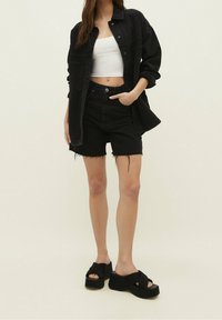 Camisa de mezclilla negra de corte holgado y con bolsillos, combinada con pantalones cortos de mezclilla negra de talle alto con bordes deshilachados, y sandalias de plataforma negra gruesa.