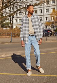 Modelo con una camisa de cuadros gris y negro, camiseta beige, jeans de azul claro y zapatos beige con cordones naranjas, de pie en una calle.