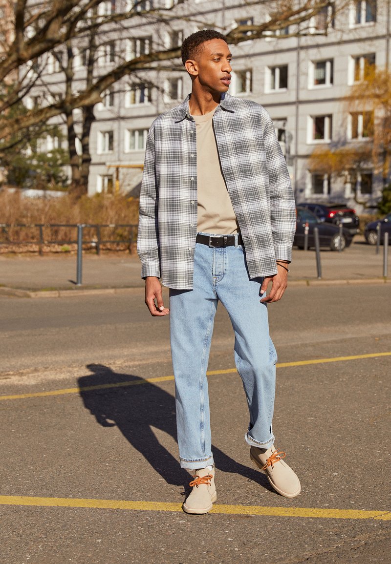 Grau-weiß kariertes Hemd über einem beigen T-Shirt, hellblaue Jeans und beigefarbene Schuhe mit orangen Schnürsenkeln. Außenbereich mit Asphaltboden.
