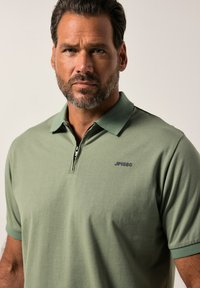 Grön polo tröja i mjukt tyg, med dragkedja och korta ärmar. "JP1880"-logotyp i mörkblått på vänster bröst.