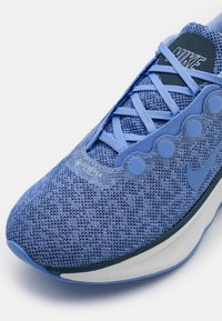 Zili Nike sporta apavi ar sietiņu augšdaļu, teksturētu apdari un izceltiem caurumiem. Iekļauti pelēki akcenti un logo uz mēles. Viegls dizains.
