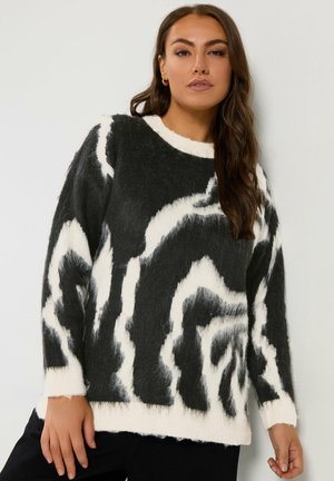 Femme portant un pull à motif duveteux noir et blanc, debout devant un fond clair uni, regardant vers l'avant avec une expression détendue.