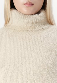 Close-up van een persoon in een beige fuzzy gebreide turtleneck trui, waarbij het onderste gedeelte van het gezicht en de hals zichtbaar is.