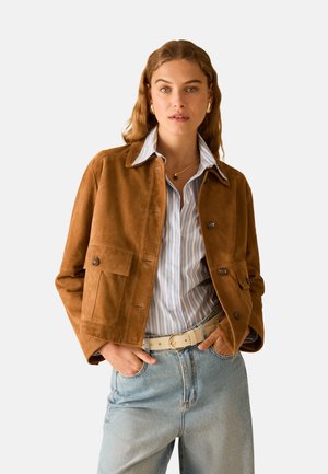 Donna che indossa una giacca in camoscio marrone sopra una camicia a righe blu, jeans azzurri a vita alta e una cintura bianca, in piedi con le mani nelle tasche.