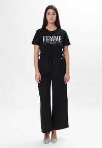Sort t-shirt med "FEMME FATALE" print, parret med sorte brede bukser. Modellen bærer sølvglitrende højhælede sko. Enkel og afslappet outfit.