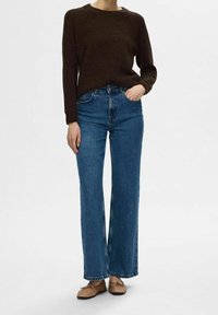 Dunkelbrauner Pullover, lockere Passform, Rundhalsausschnitt; hochtaillierte blaue Jeans, weite Beinform; beige Ballerinas mit Schleifendetail; vor einem einfarbigen Hintergrund.