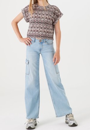 Kvinde iført en mønstret bluse med korte ærmer, lyseblå vidde-ben cargo-jegns og grå sneakers, stående med den ene hånd på hoften.