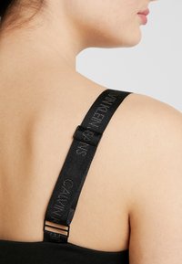 Cintura nera con testo in rilievo, hardware in metallo regolabile, attaccata a un tessuto scuro. Il tono della pelle è visibile sulla spalla.