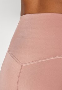 Leggings taille haute en tissu doux et extensible, présentant une texture lisse et une couleur rose pâle avec des détails de couture subtils.
