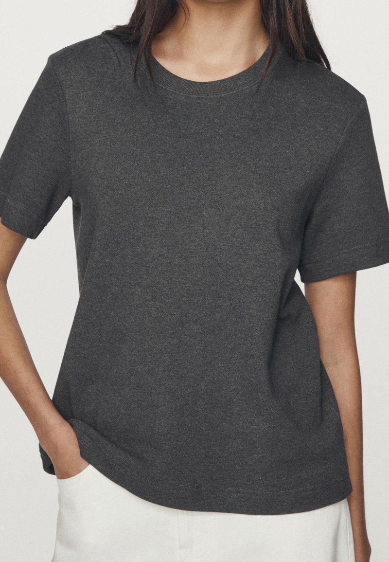 Femme portant un t-shirt gris foncé à manches courtes avec un col rond et un pantalon blanc, main dans la poche, sur un fond uni.