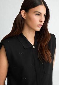 Mustmata ilma varrukat blouse kaelusega, millel on kuldne nööppealne ja mustad dekoratiivsed detailid. Sile, kerge kangas, millel on peen sära.