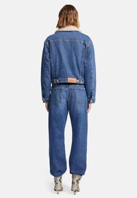 Jeansowa kurtka z owczym kołnierzem, w niebieskim odcieniu, o dopasowanym kroju. Zestawiona z luźnymi niebieskimi spodniami jeansowymi i wzorzystymi botkami do kostki.