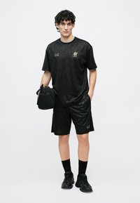 Melnas sporta apģērbs ar rakstainu īsroku kreklu un šortiem, papildināts ar melnu somu un atbilstošiem apaviem. Redzami zelta akcenti.