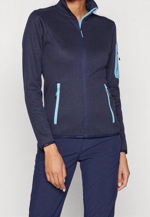 Vrouw draagt een nauwsluitend marineblauw jack met rits en lichtblauwe ritssluitingen op de zakken en mouw, gecombineerd met marineblauwe broek.