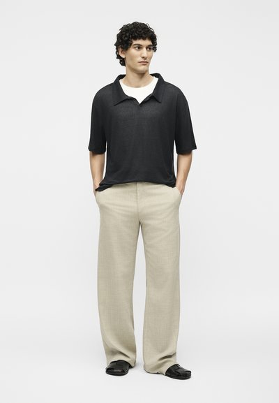 Άνδρας με σγουρά μαλλιά που φοράει μαύρο κοντομάνικο πουκάμισο με γιακά, μπεζ φαρδύ παντελόνι και μαύρα σανδάλια, στέκεται με τα χέρια στις τσέπες.