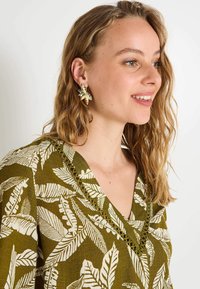 Haut en textile vert avec un motif de feuilles blanches, présentant un décolleté en V et une couture décorative. Modèle portant des boucles d'oreilles dorées florales. Cheveux longs et ondulés.