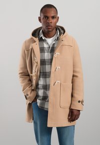 Beige duffelrock i ullblandning med knappar, huva och sidofickor, bärs över en rutmönstrad skjorta och blå jeans.