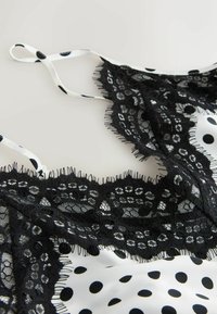 Un borde de encaje negro con flecos adorna una tela blanca con lunares negros; tirantes finos complementan el diseño delicado.