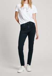 Tmavě modré skinny džíny vyrobené z denimu, kombinované s bílým tričkem s metalickým grafickým textem. Nosí se s bílými teniskami.