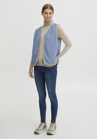 Fleecasti prsluk svijetloplave boje s patentnim zatvaračem, nošen preko bež turtleneck majice. Uparen s plavim uskim trapericama i sivim sportskim cipelama.