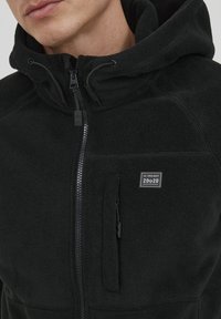 Black fleece jacket with a zip front, adjustable hood, and side pocket. Features a small logo patch on the chest. Soft texture.
